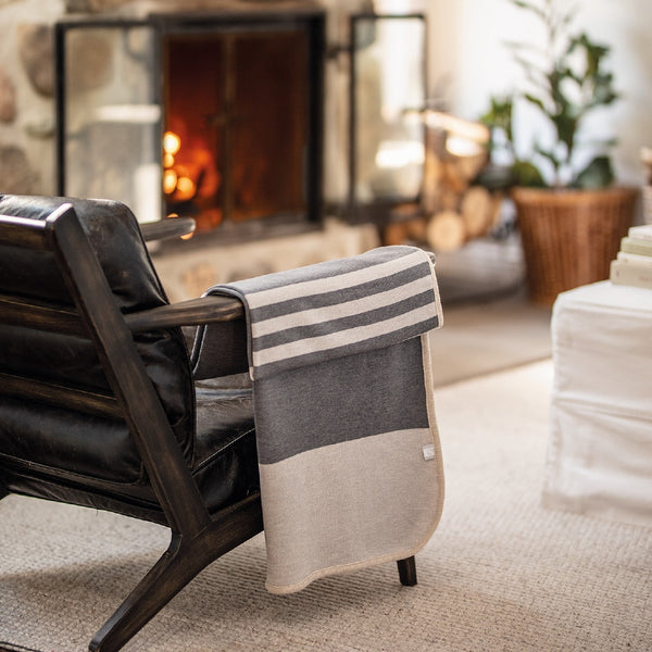 Jeté gris avec rayures blanches en laine de mérinos devant foyer / Grey with white stripes merino wool throw in front of fire place made in Canada by Volprivé.