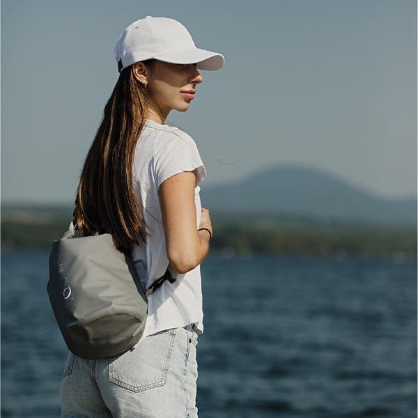 Jeune femme sur un quai avec lac et montagne et sac au sec 5L gris par Volprivé / Young women with white cap on dock with water background and 5L dry bag by Volprivé.