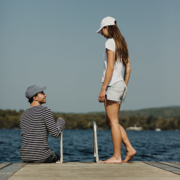 Jeune couple sur un quai bord de lac avec casquettes bleu et blanche fabriqués par Volprivé 100% coton / Couple sitting on a dock of a bay near lake wearing blue and white cap made in Canada by Volprivé.