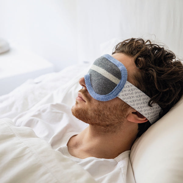 Jeune homme couché dans un lit blanc avec masque de sommeil lesté gris et bleu pâle ajustable / Young man sleeping in white bed with weighted sleep mask in grey and light blue made by Volprivé.