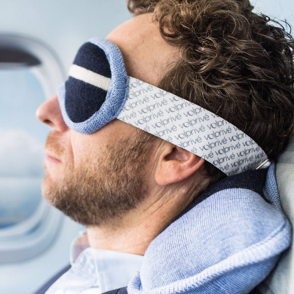 Homme portant masque de sommeil lesté bleu marine et bleu pâle en avion siège hublot / Man wearing a navy blue and light blue weighted sleep mask on plane window seat made by Volprivé.