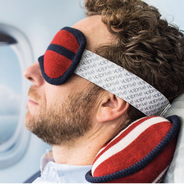 Homme assis côté hublot avec masque et oreiller rouge / Business man sitting in a plane wearing a sleep mask and travel pillow red and navy by Volprivé.