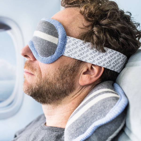 Homme assis côté hublot avec masque et oreiller gris et bleu pâle / Man sitting in a plane wearing a sleep mask and travel pillow grey and light blue by Volprivé.