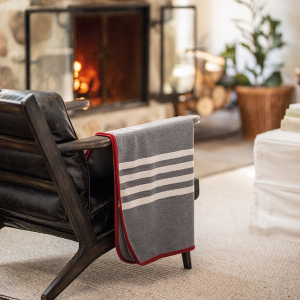 Jeté gris avec rayures blanches et contour rouge en laine de mérinos devant foyer / Grey with white stripes and red contour in merino wool throw in front of fire place made in Canada by Volprivé.